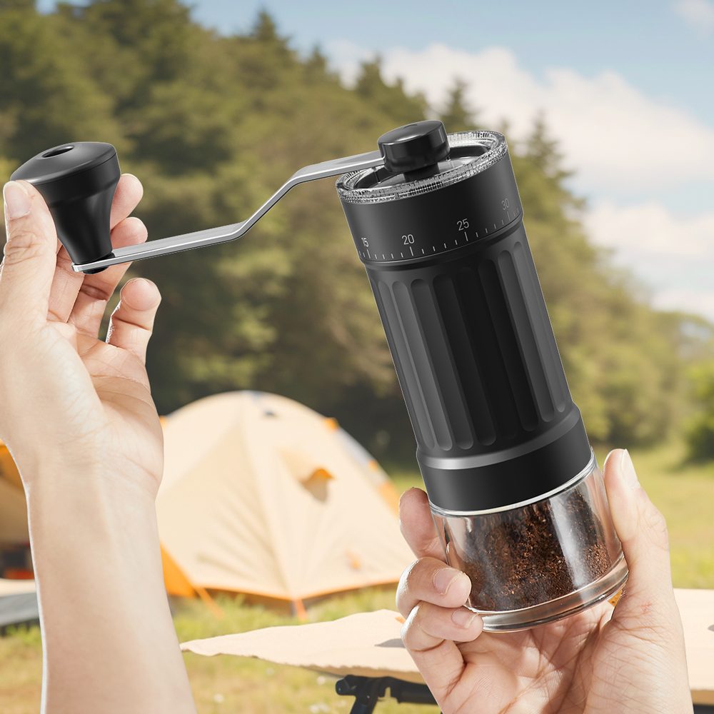 Two hands holding the Ermona hand coffee grinder outdoors, its glass jar full of fresh grounds, tents behind.