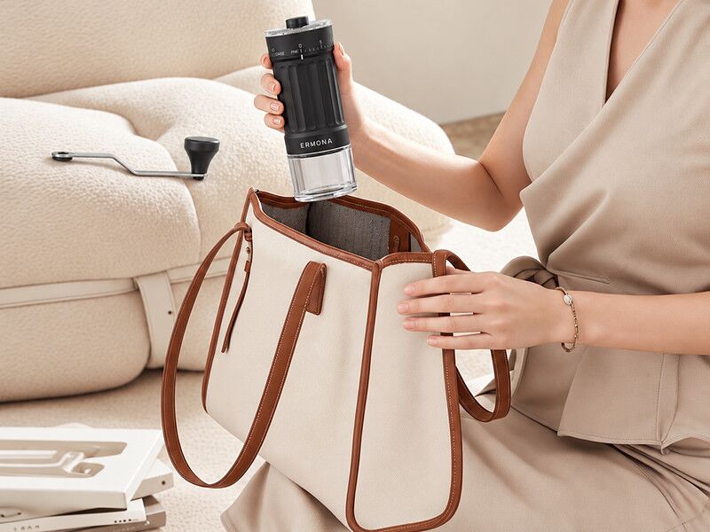 A woman packing the Ermona manual coffee grinder into a tote bag, with the detached crank resting on the sofa beside her.
